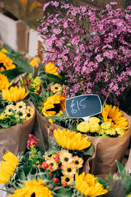 A vibrant display of fresh floral bouquets arranged with bright yellow sunflowers, small white and yellow daisies, and red and pink accent flowers wrapped in brown paper, set against a backdrop of lus