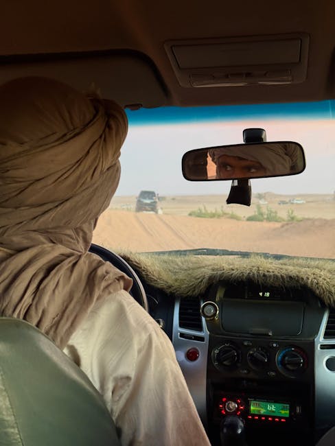 The image shows the interior of a vehicle with a woman sitting in the front passenger seat, wearing a beige hijab. The dashboard is visible, with controls and a digital display, and the rearview mirro