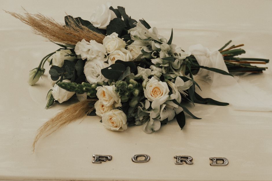 A bouquet of fresh flowers resting on a light beige surface, featuring white roses, white lisianthus, green foliage, and some dried beige pampas grass. The bouquet is arranged thoughtfully with the fl