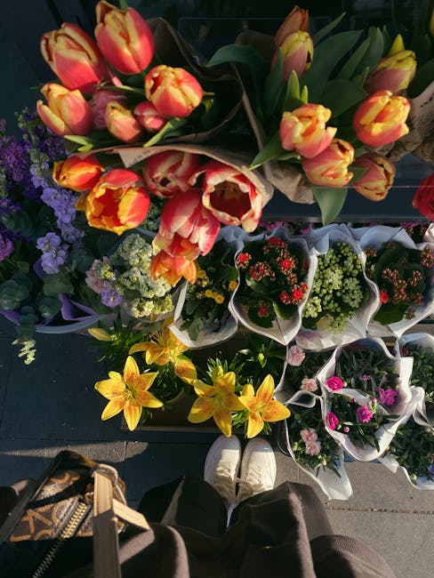 A display of vibrant flower arrangements at a market stall showing a variety of fresh blooms. The top bouquet features orange and yellow tulips with hints of red, arranged in a natural style with gree